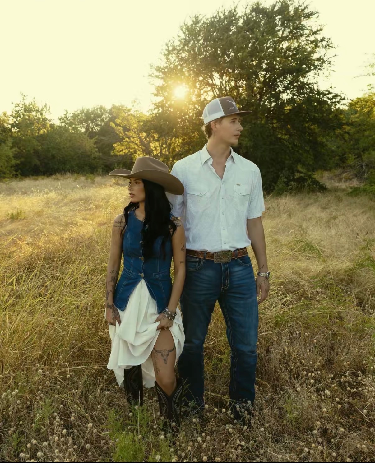 Couple in Western wear standing in tall golden grass at sunset, she in a felt hat and denim vest, he in a pearl-snap shirt and trucker cap